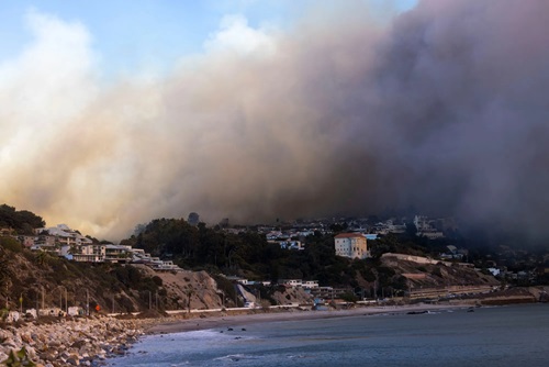 A coastal neighborhood on a hillside under a massive wall of thick, dark wildfire smoke looming over houses, trees, and the ocean shore below.