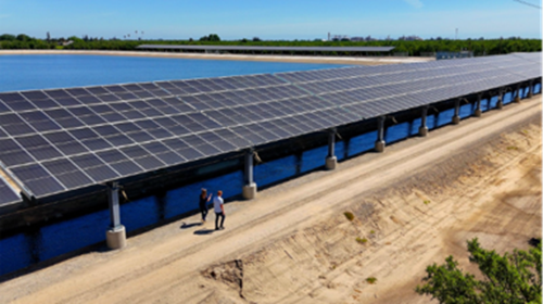 Project Nexus in California’s Turlock Irrigation District, showing rows of solar panels installed above an irrigation canal. Two people walk along the canal access road beneath the panels, with the blue water visible below and agricultural fields and an industrial skyline in the distance