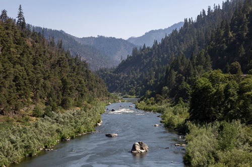 The Klamath River flows through a dense, forest-covered canyon, with large rocks sticking out of the blue-gray water under a hazy sky.