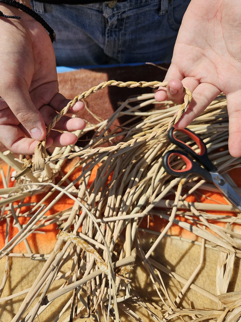 Hands weaving a braided cord from dried plant fibers beside a pair of scissors and weaving materials.