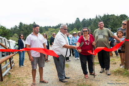 Ribbon-cutting ceremony outdoors with a group of people as two participants cut a red ribbon with oversized scissors. Photo by Kellie Brown.