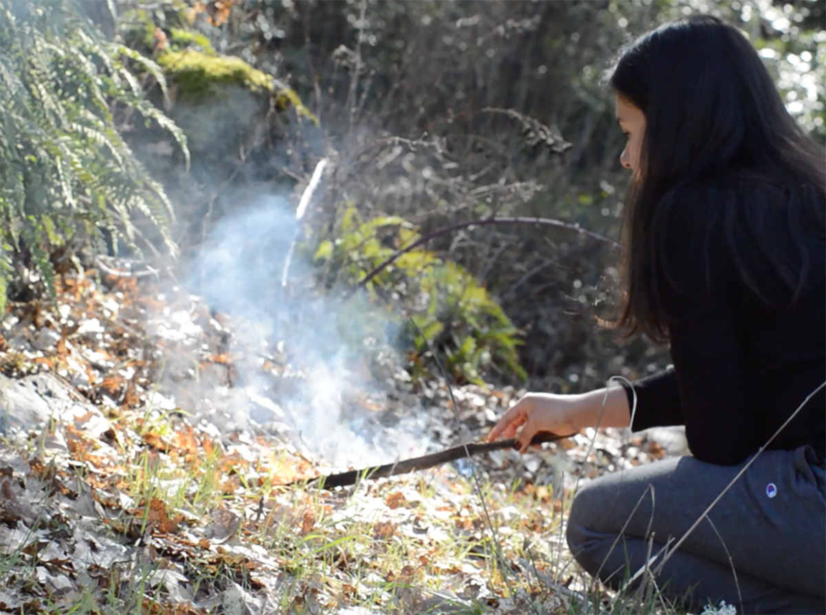 Person crouching in a forested area, using a stick to tend a small, smoky ground fire among dry leaves and grass.