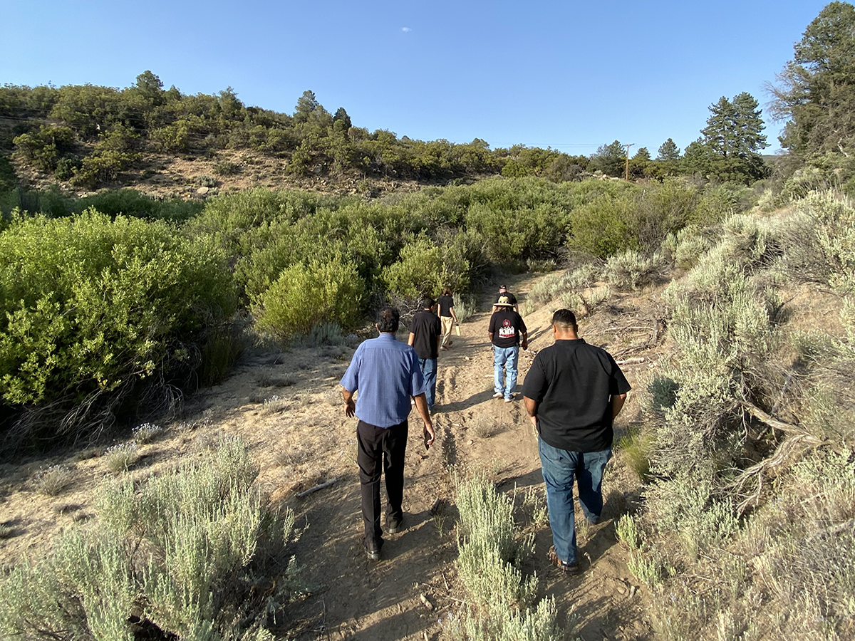 Group of people walking up a dirt trail through dry, shrub-covered hills under a clear blue sky.